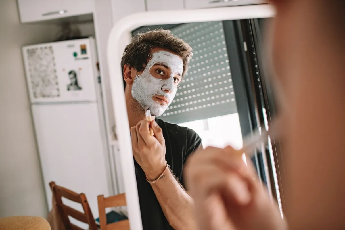 Man applying a facial mask while looking in a mirror, illustrating skincare routines.
