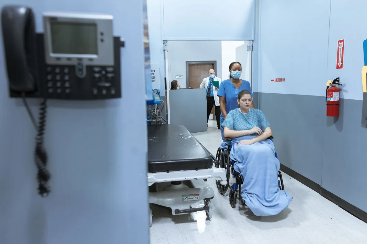 Patient in a wheelchair being assisted by a healthcare worker in a hospital corridor.