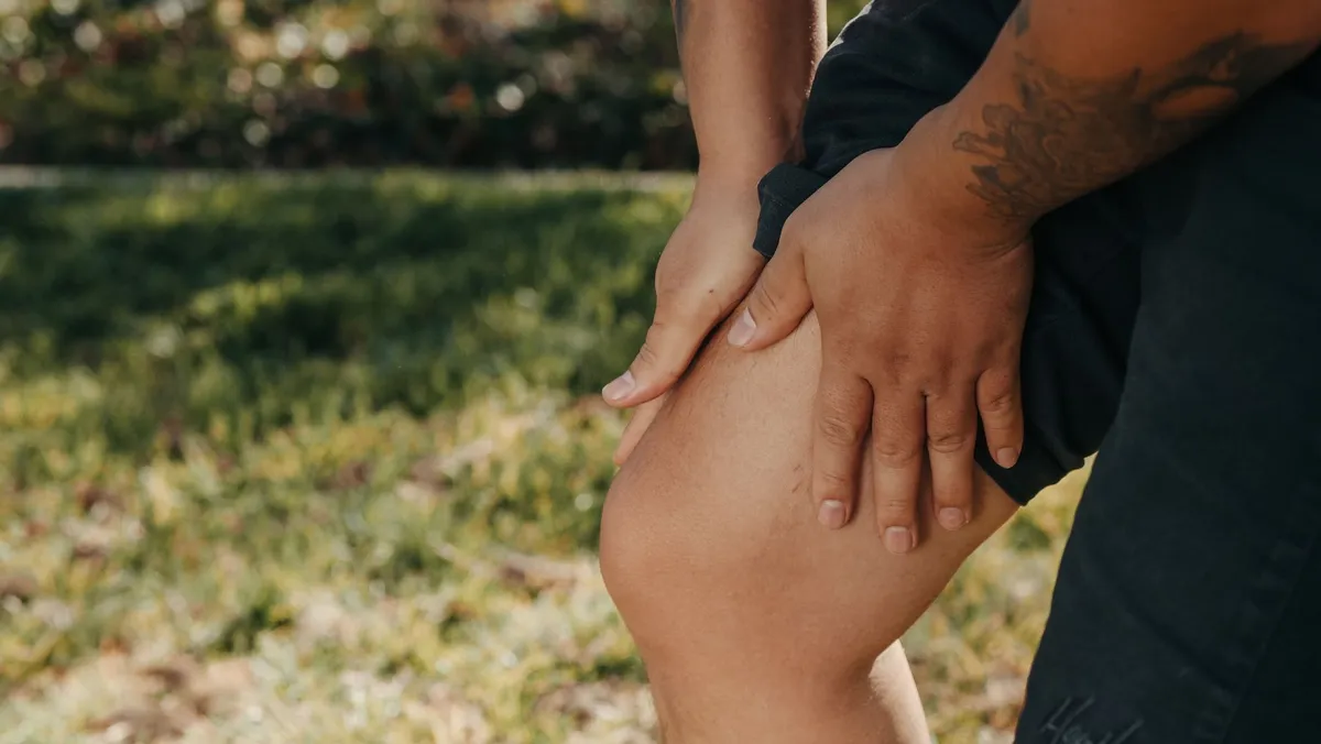 Person holding their knee while sitting on grass, indicating potential discomfort or injury.