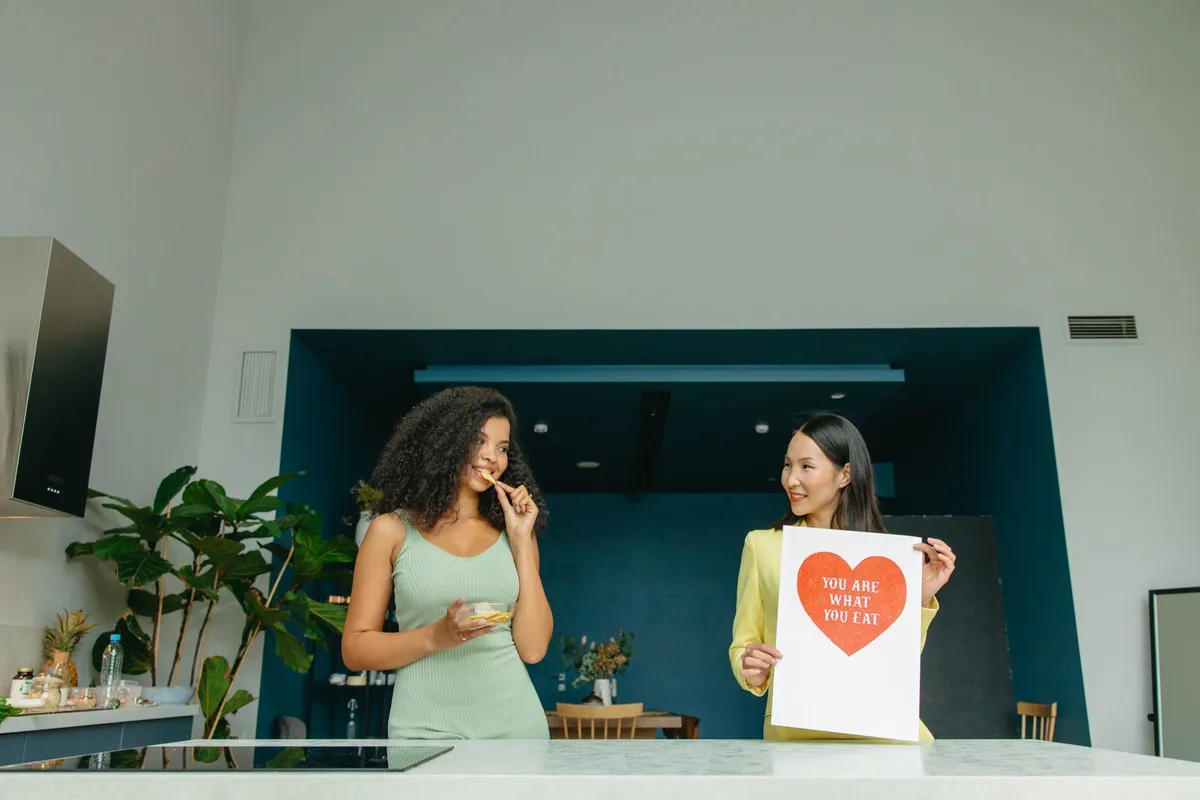 Two women discussing nutrition in a modern kitchen, one holding a sign about healthy eating.