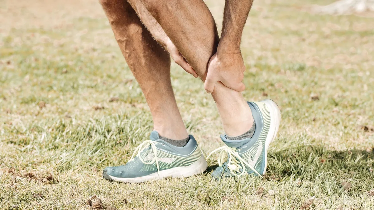 Person in athletic shoes holding their ankle in a grassy area, indicating potential injury.