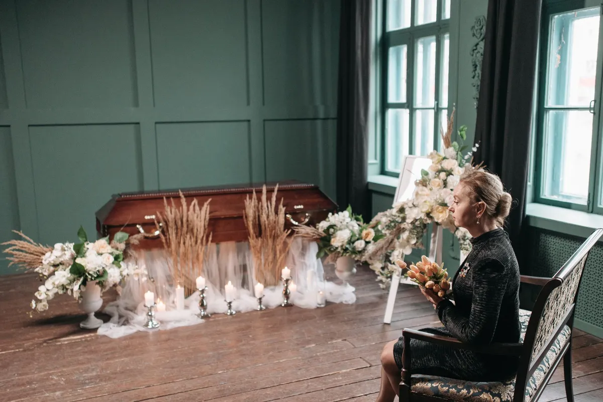 Woman sitting in front of a decorated coffin with flowers and candles in a dim room.