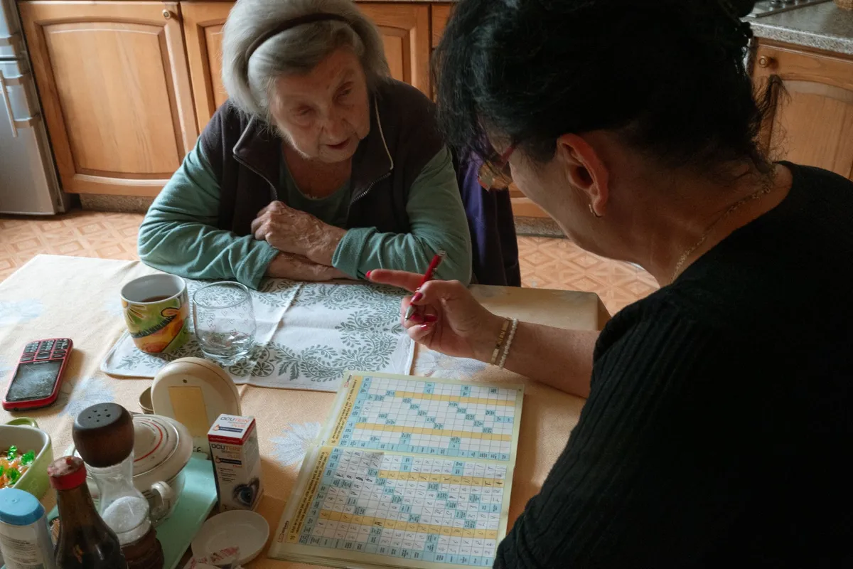 Caregiver assisting elderly woman with a puzzle in a cozy kitchen setting.
