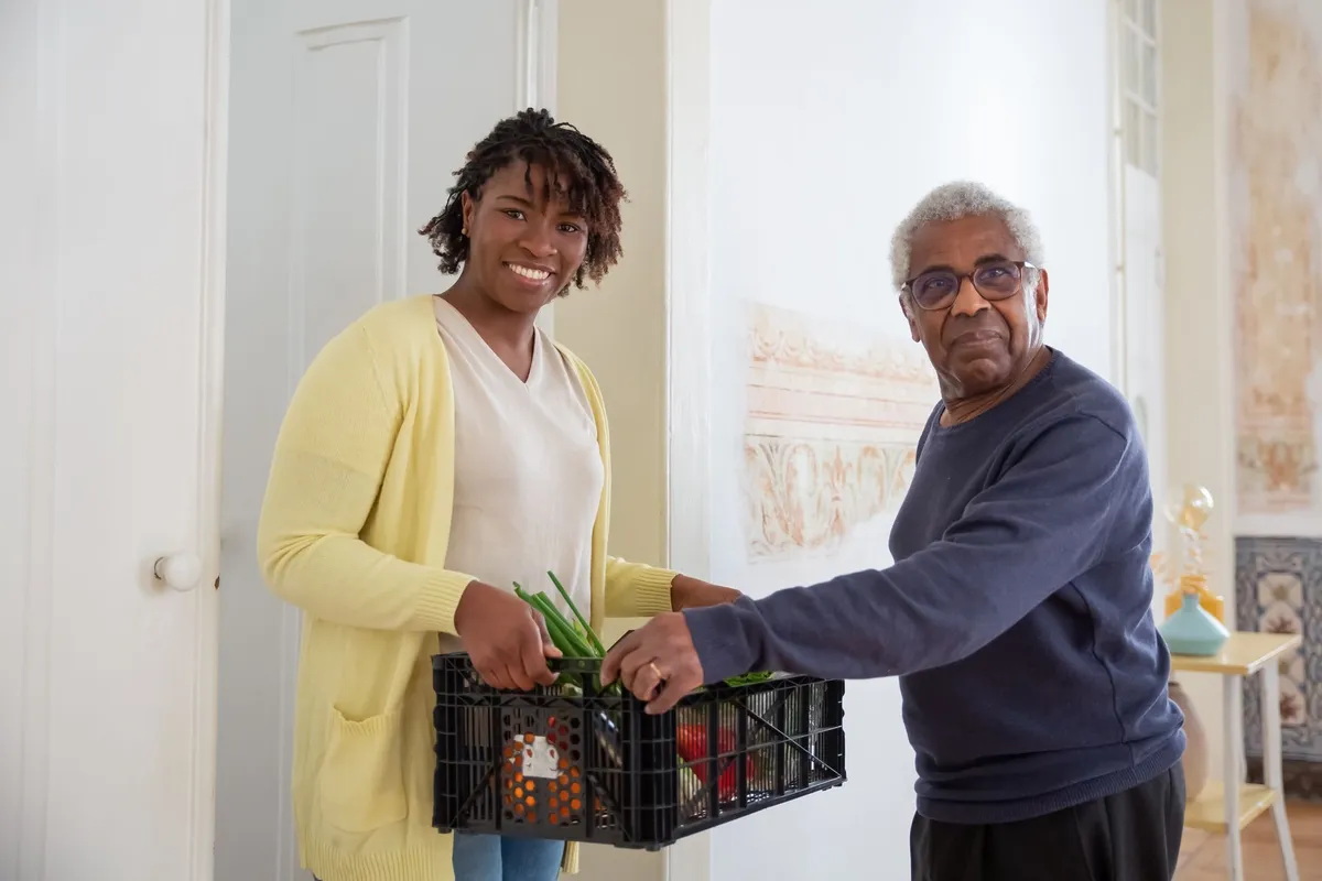 Caregiver and elderly man holding a crate of vegetables indoors