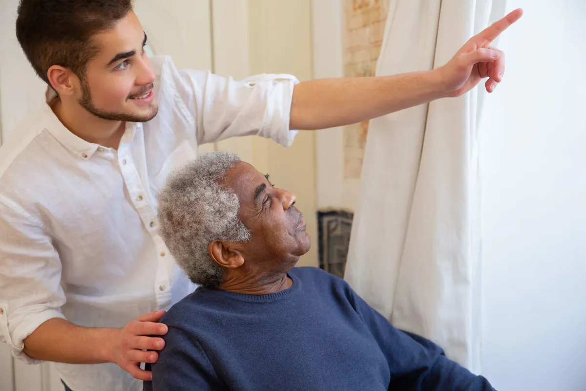 A young man points out the window while assisting an older man in a chair.
