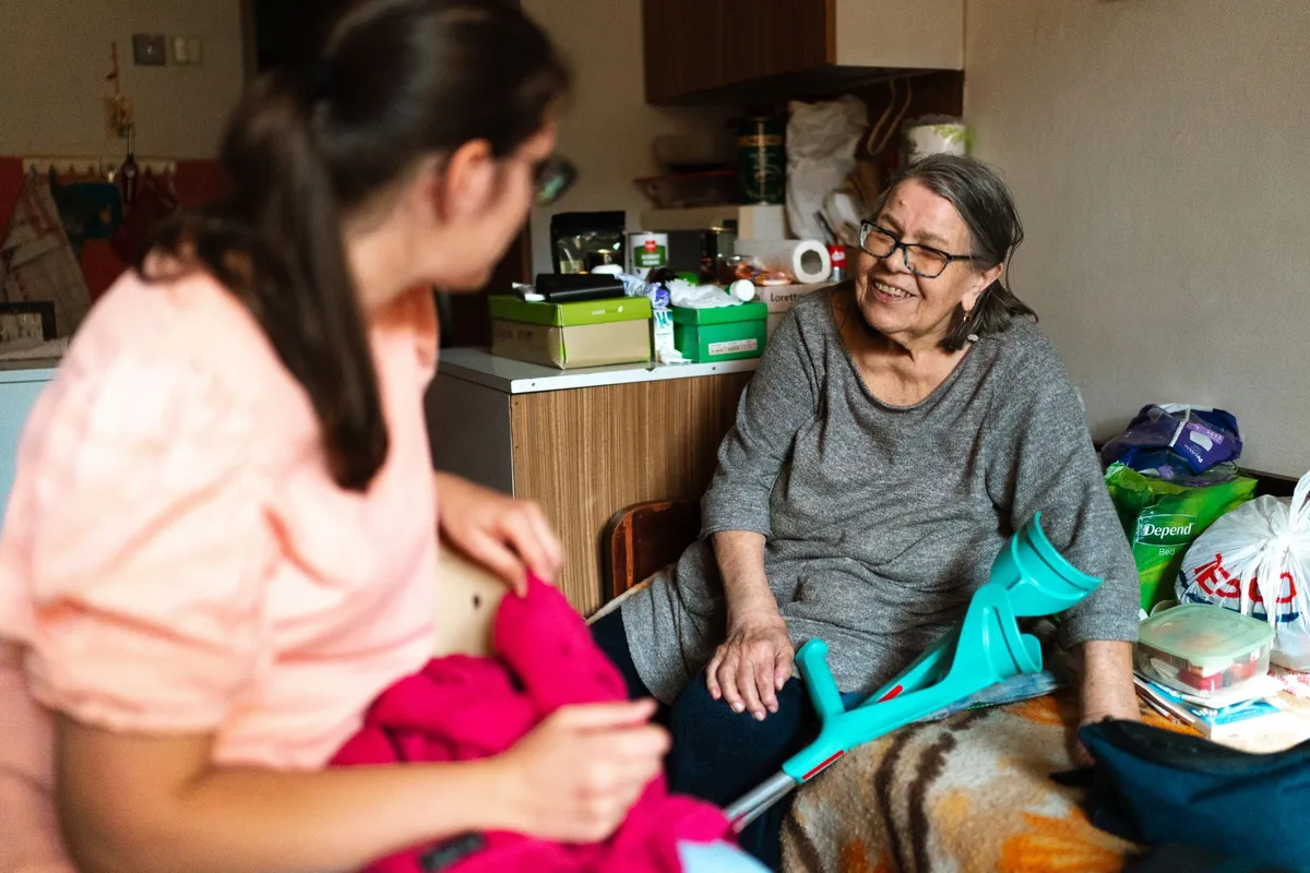 A caregiver and an elderly woman smiling and talking in a cozy room.