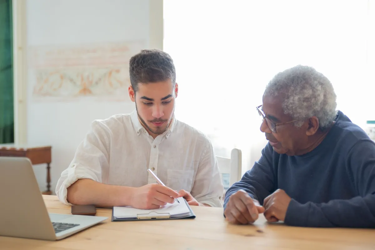 Young man assisting elderly man with paperwork at a table with a laptop.