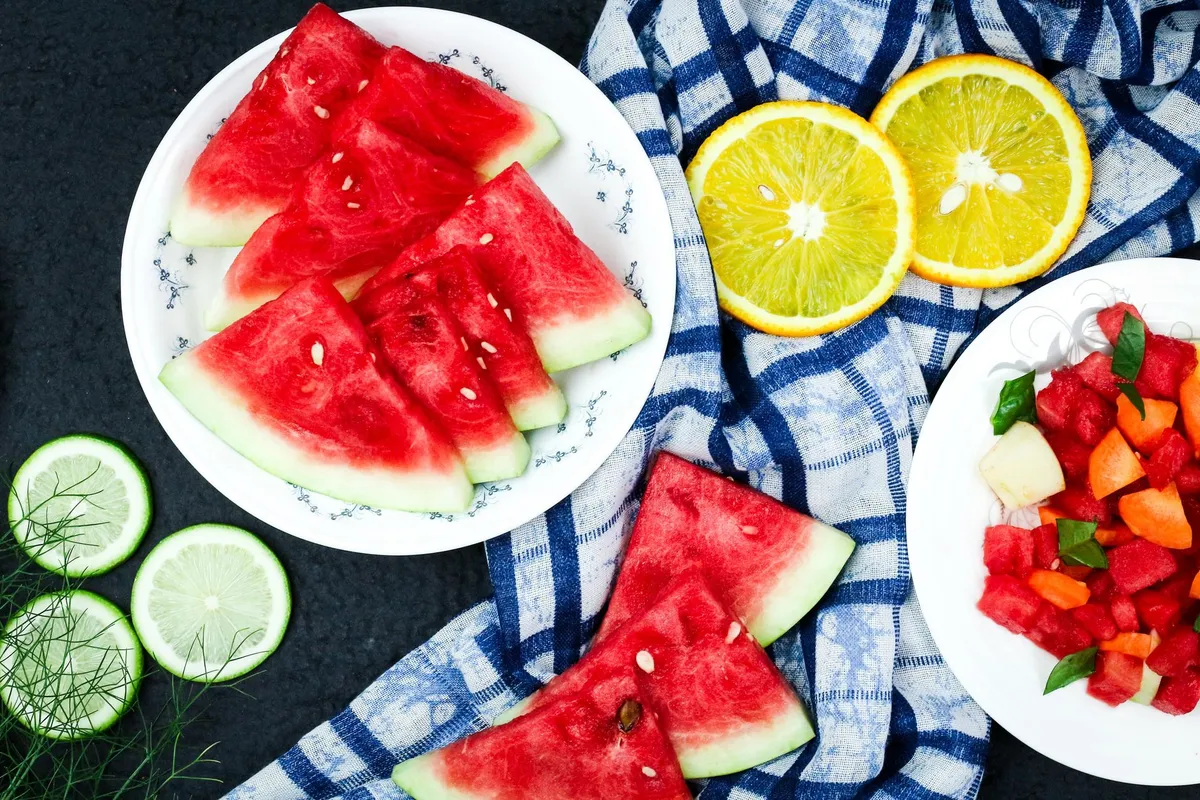 Sliced watermelon and oranges on a blue checkered cloth with a plate of mixed fruit.